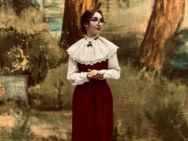 EstelaYake with glasses, dark hair in a bun, wearing a red dress and white lace collar, standing with hands together, looking to the side, in a vintage-style photo.