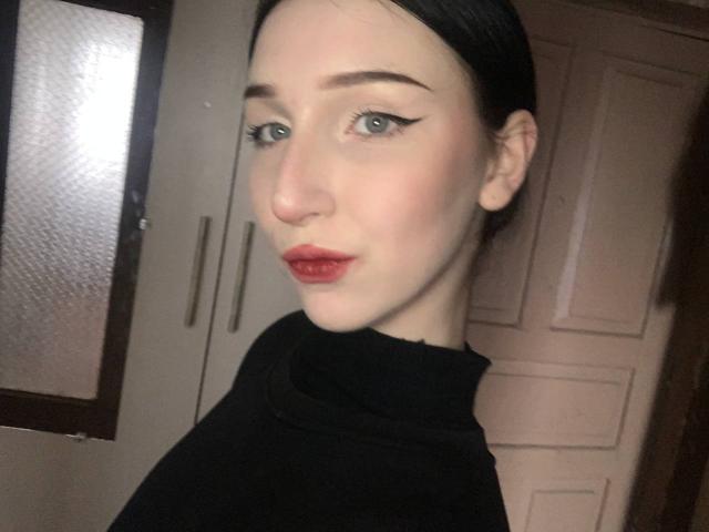 EstelaYake has pale skin, dark hair, blue eyes, and red lipstick with black winged eyeliner. She's wearing a black turtleneck, looking at the camera with a slight pout.