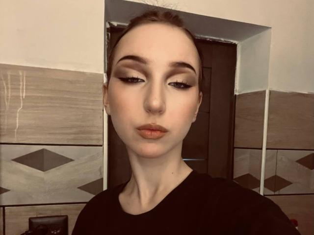 EstelaYake has pale skin, dark eyeliner, and brown lips. She's wearing a black shirt, with her brown hair pulled back, looking serious. Her tits are small.