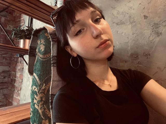 EstelaYake with short black hair, small hoop earrings, black shirt, subtle makeup, and a relaxed expression, sitting in a chair with green and gold patterned fabric.