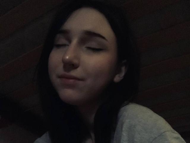 EstelaYake with dark hair, closed eyes, and slight smile, wearing a gray shirt, has a small nose ring, and looks relaxed in dim light.