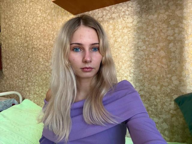 SaraiCartmill with blue eyes, blonde hair, and a purple off-shoulder top, looking at the camera with a neutral expression, sitting in a room with textured wallpaper.
