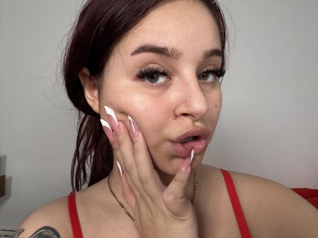 Luucie with red hair, pouty lips, and long pink nails touching her cheek. She's wearing a red top, has green eyes, and a tattoo on her left shoulder.