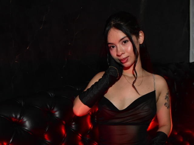 MelodyGloss with black gloves, sheer black dress, and tattoo on her right shoulder, sitting on a tufted black leather couch, looking at the camera with a slight smile.