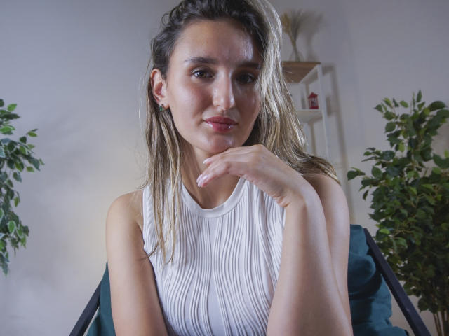 Kelly Cowling mit blonden Haaren, einem weißen, gerippten Top ohne Ärmel, leicht feuchten Haaren, der Hand in der Nähe des Kinns, einer grünen Pflanze links und einem weißen Regal rechts, sitzt auf einem blauen Stuhl und schaut in die Kamera..