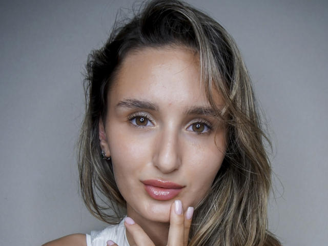 Kelly Cowling hat braune Augen, glänzende rosa Lippen und leicht zerzaustes blondes Haar. Sie berührt ihr Kinn mit den Fingern und zeigt dabei ihren rosa Nagellack..