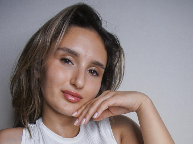 KellyCowling with medium brown hair, light skin, and glossy pink lips, rests her hand on her cheek, wearing a white sleeveless top, looking slightly to the side.