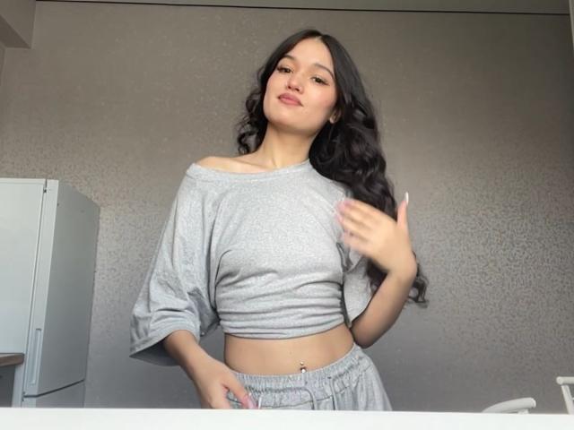 Shaquita Horwich, avec de longs cheveux noirs ondulés, portant un haut court gris à une épaule et un pantalon gris assorti, une main sur la hanche, l'autre main levée, un léger sourire, vernis à ongles rose..