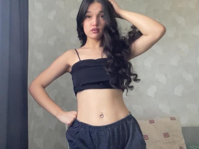 Shaquita Horwich, avec ses longs cheveux noirs et bouclés, porte un haut noir sans bretelles qui expose ses petites seins et son piercing au nombril. Elle porte un pantalon noir ample et se tient debout, une main sur la tête, en regardant l'appareil photo..