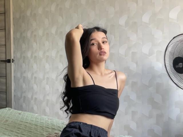 ShaquitaHorwich with long black hair, black strapless top, and dark pants, pouting with arms raised, standing in a room with a green bed and a fan.