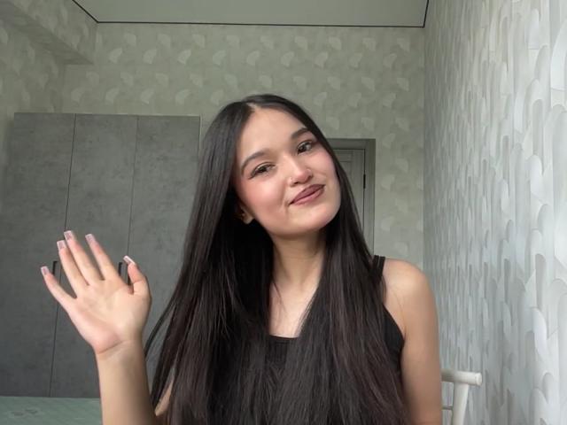Shaquita Horwich, avec ses longs cheveux noirs, souriante et agitant la main ornée d'ongles roses, porte un haut noir sans manches, dans une pièce aux motifs blancs. Elle a un joli visage, un petit nez et de douces lèvres roses..