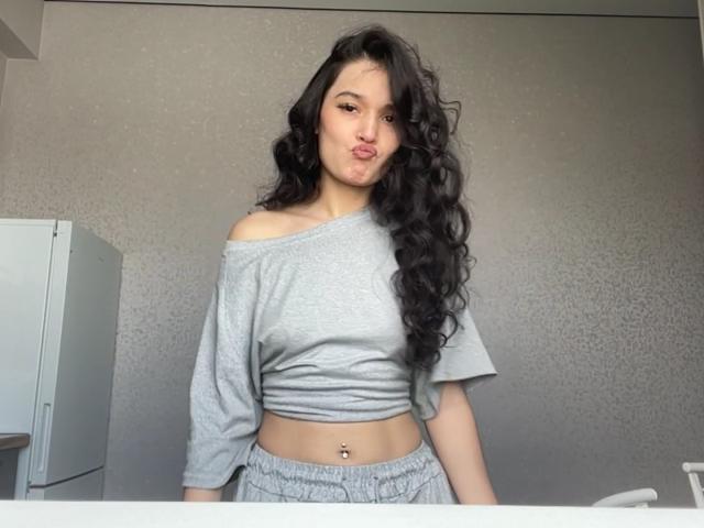 Shaquita Horwich, avec ses longs cheveux noirs bouclés, son haut court gris à épaules dénudées, qui révèle de petites seins, et son pantalon gris, a un piercing au nombril et poute ses lèvres..