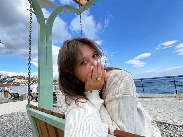 PauletteRylander with brown hair, white sweater, and ringed hand, covering her mouth, sitting on a green swing by the blue sea, under a clear sky.