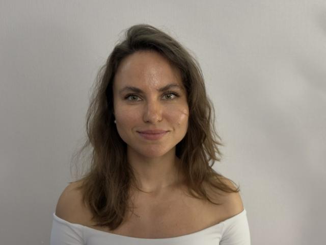 TangelaEmmel has wavy brown hair, green eyes, and a slight smile. She's wearing an off-shoulder white top, showing her bare shoulders and upper chest.