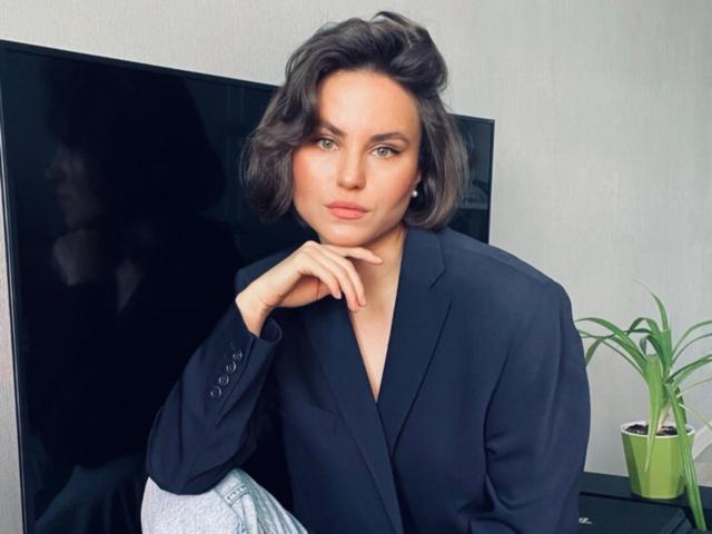 TangelaEmmel with short, wavy dark hair, green eyes, and pink lips, wearing a black blazer, hand on chin, pensive expression, sitting next to a green plant.