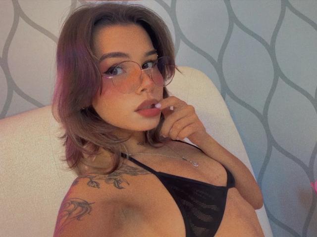 Lena, avec des lunettes, cheveux bruns, soutien-gorge en dentelle noire, tatouage sur l'épaule, doigt dans la bouche, regardant vers le haut, rouge à lèvres rose clair, sourire discret, détendue, sexy, éclairage doux..