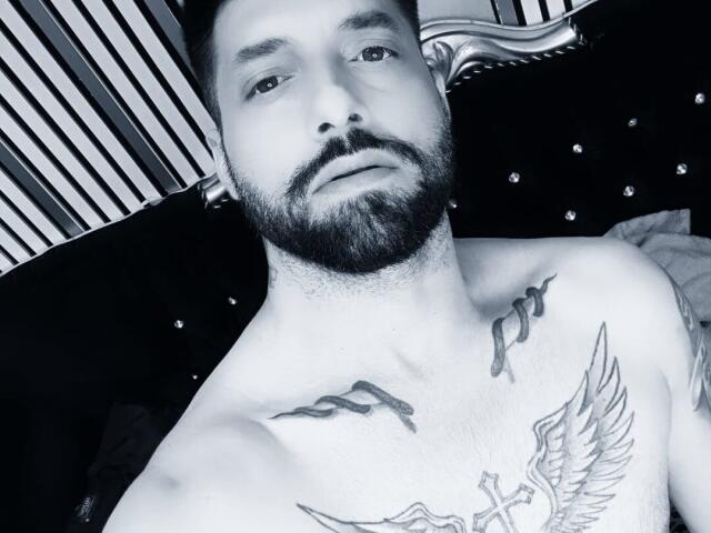 TommyDum, shirtless with a beard, has tattoos on his chest including a bird and wings, and is lying on a black tufted bed with striped pillows.