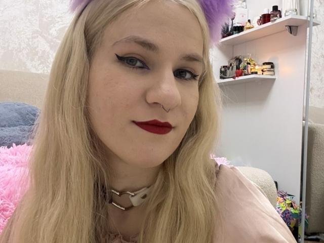 AlejandraBobo with pale skin, blonde hair, purple cat ears, red lipstick, black eyeliner, nose ring, choker, pink fluffy pillow, and a cute, slightly nerdy, sexy look.
