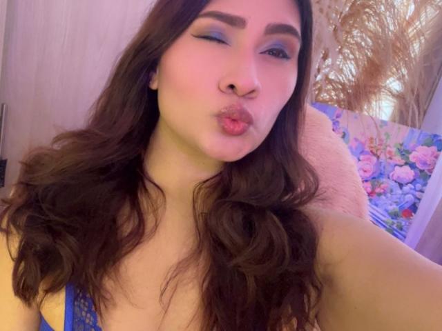 ElyRoss with long brown hair, blue eyeliner, and pink lipstick, puckering her lips in a selfie, wearing a blue lace top, showing her tits slightly.