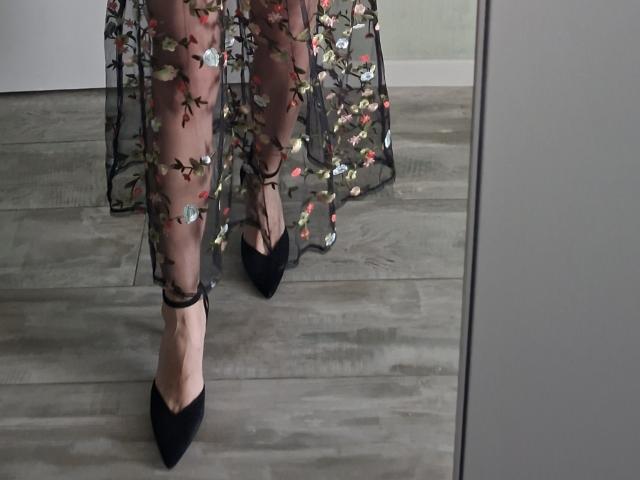 CrystalHope's legs wearing black pointed heels, sheer floral-patterned black dress, standing on grey wooden floor, visible reflection in mirror.