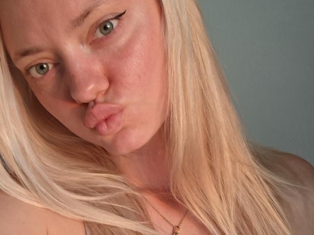 CrystalHope with pouty pink lips, blonde hair, and green eyes, making a kissing face. She has a small nose and wears a thin gold necklace. Her tits are not visible.