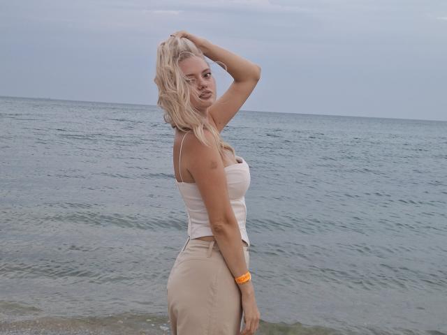 CrystalHope, blonde, wavy hair, white tank top, tight beige skirt, orange wristband, hand in hair, standing at beach, ocean behind, gray sky, curvy, large boobs, confident, sexy.