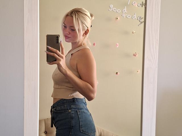 CrystalHope with blonde hair in a bun, wearing a beige tank top and blue jeans, taking a mirror selfie with a smartphone, showing her ass and sideboob.