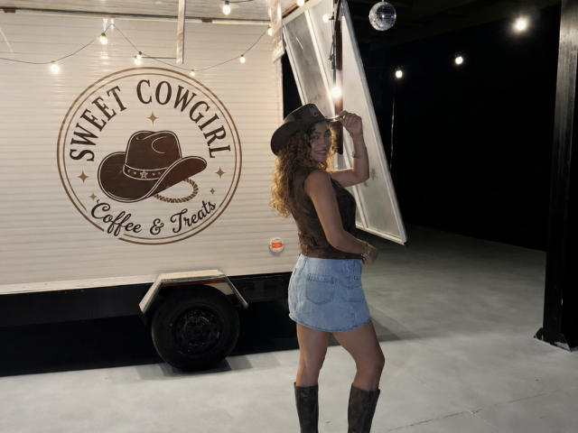AriannaBlake with curly brown hair, wearing a brown cowboy hat, brown sleeveless top, denim skirt, and brown boots, lifting a trailer door, in a dimly lit garage.