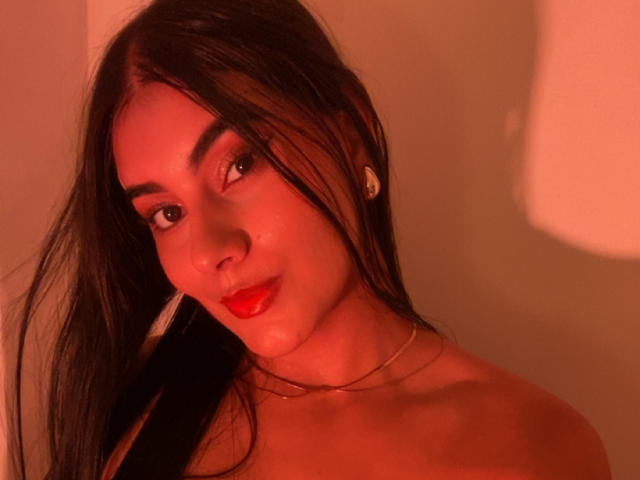 AlaraKey with long black hair, red lipstick, and red light, wearing a thin gold necklace, looking at the camera, showing bare shoulders, slight smile, small gold earrings.