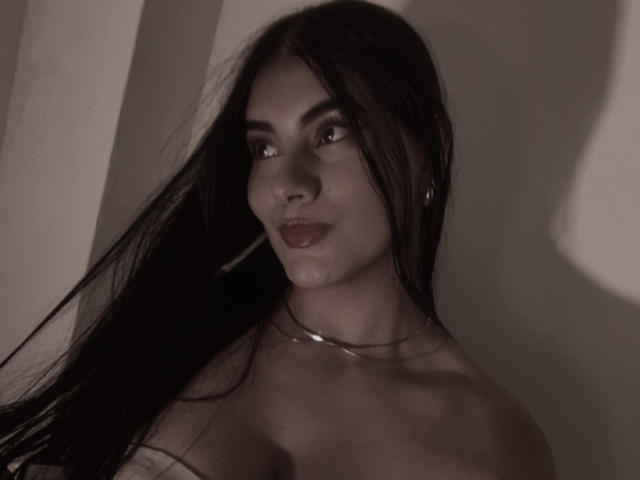AlaraKey has long black hair, brown eyes, and is smiling with her lips slightly parted. She's topless with a thin necklace and her tits are visible. Dark, moody photo.