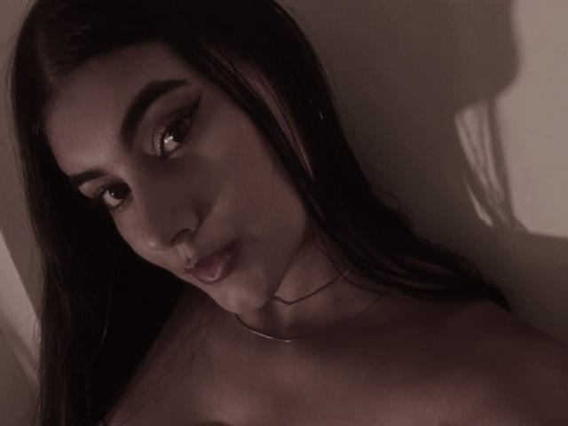 AlaraKey has dark brown hair, thick eyebrows, and is looking slightly to the side with her lips slightly parted. Her bare shoulders are visible. Dim, shadowy lighting.