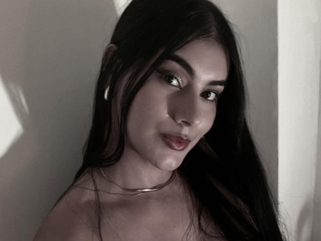 AlaraKey has long black hair, dark eyes, and a slight smile. She's wearing a silver choker and has a shiny, wet-looking nose. The photo is in grayscale.