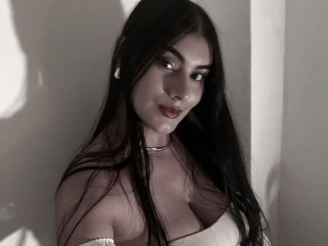 AlaraKey with long black hair, wearing a white off-shoulder top, showing her big boobs, dark eyes, and a subtle smile, in a soft-lit, grayscale photo.