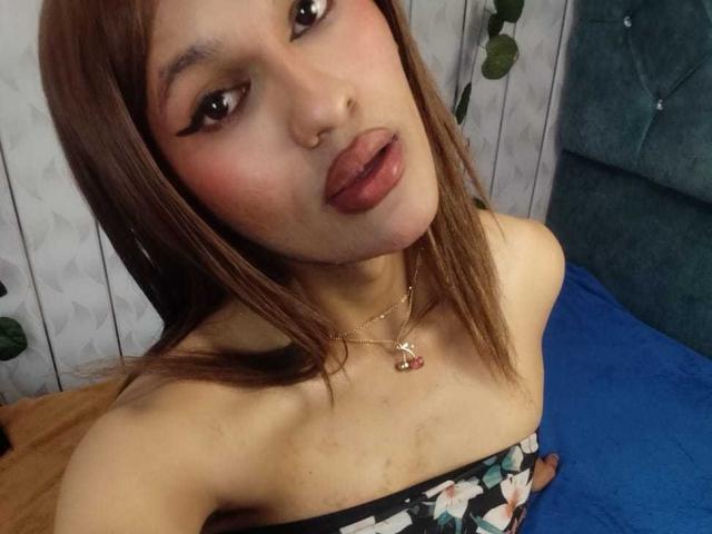 PamelaXFox with brown hair, pink eyeliner, and pouty lips, wearing a floral strapless top, necklace, and pink nose ring, looking up at the camera.