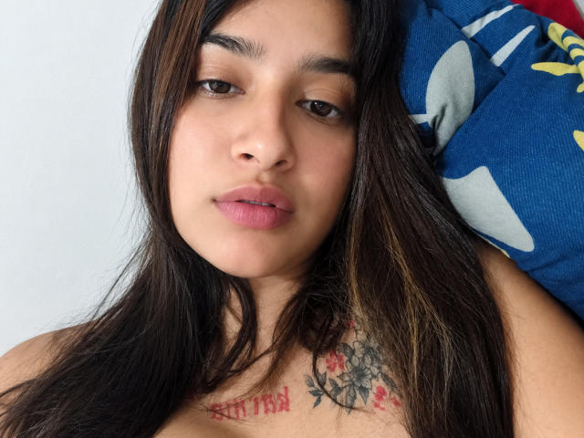SofiTattoo has tan skin, brown eyes, and long dark brown hair with subtle highlights. She's topless, showing a tattoo of roses and text on her chest, with pink lips.