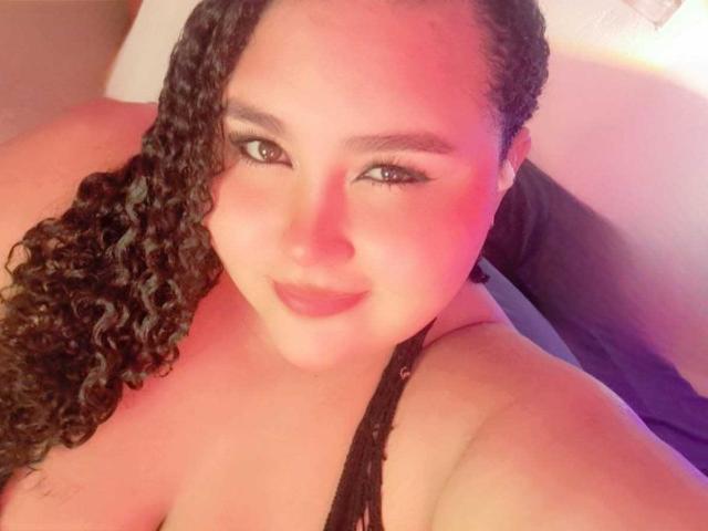 HurrenMatt has curly black hair, brown eyes, and a cute smile with pinkish-red lighting on her face. She's wearing a black strap, showing her large tits.