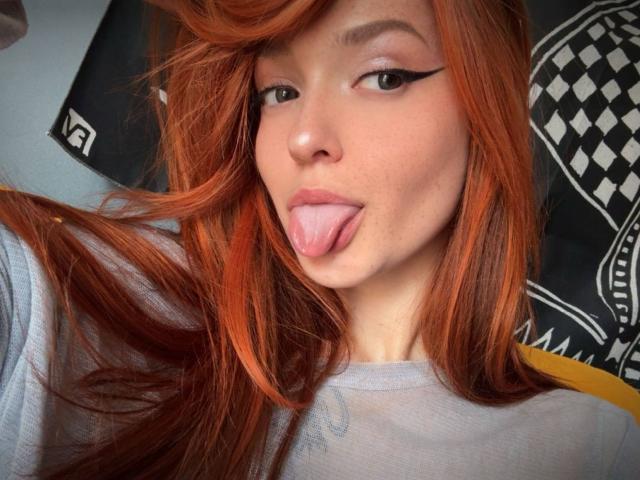 IrishReversed with red hair, black eyeliner, tongue out, freckles, and a light gray shirt, showing off her cute face and playful expression.