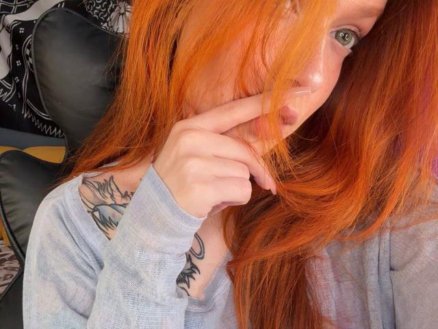 IrishReversed with bright orange hair, green eyes, and a tattoo on her chest, wearing a grey shirt, has her finger near her mouth, showing off her tits.