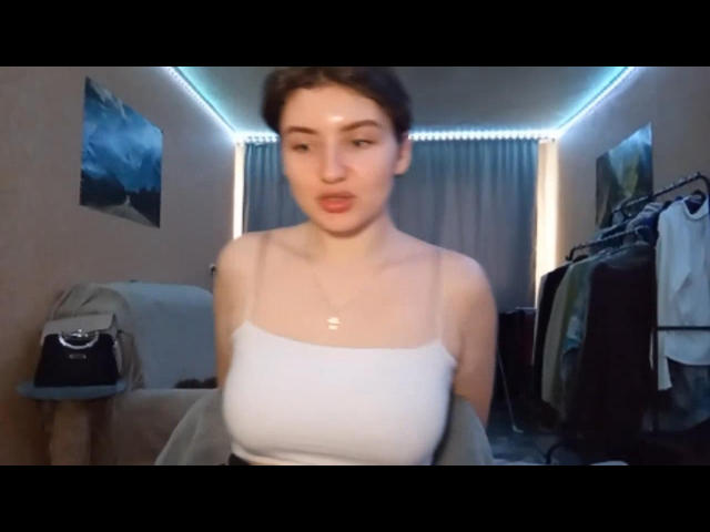 EliaPharmes has pale skin, brown hair in a bun, small tits, and a white spaghetti strap top. She's in a bedroom with clothes and a bag in the background.