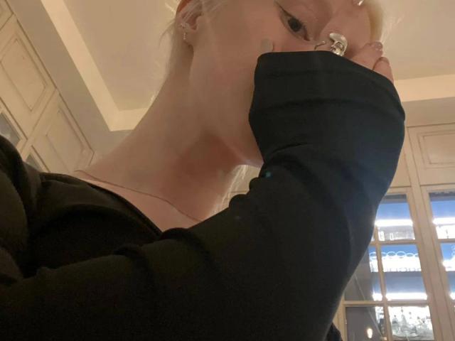 Enigmática, com cabelo loiro, uma manga preta cobrindo o nariz, um anel no dedo, blusa preta, pele clara, uma tatuagem levemente visível na bochecha, fotografada de baixo..