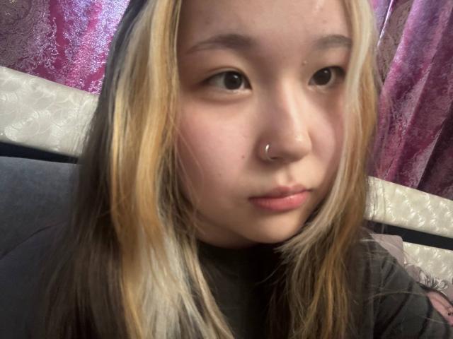 JungMcgranor has blonde and brown hair, a nose ring, and is looking at the camera with slightly parted lips, wearing a black shirt.