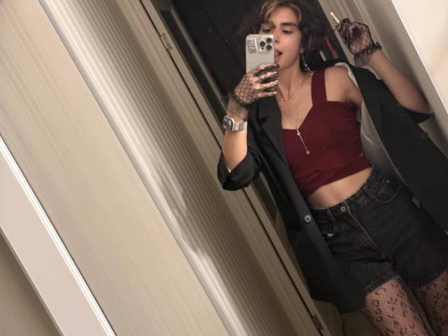 TameikaMacclellan with short brown hair, tongue out, wearing a red crop top, black jacket, high-waisted shorts, lace gloves, and patterned tights, taking a mirror selfie.