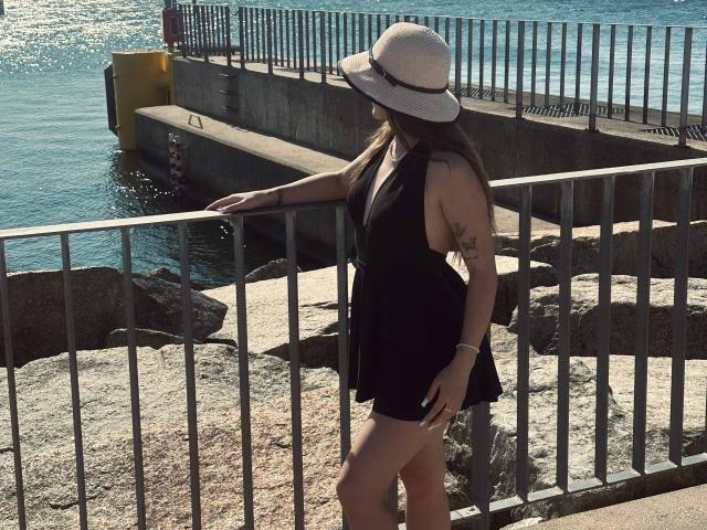 MisscharyNessy with long brown hair, wearing a white sunhat, black sleeveless dress, and tattoo on her right arm, standing by a metal railing, near a rocky waterfront.