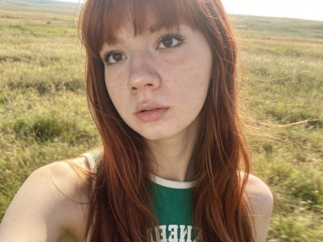 HalleyBabiracki has red hair, freckles, and green eyes, wearing a green top, with a serious expression, showing her bare shoulders and a slight pout, in a grassy field.