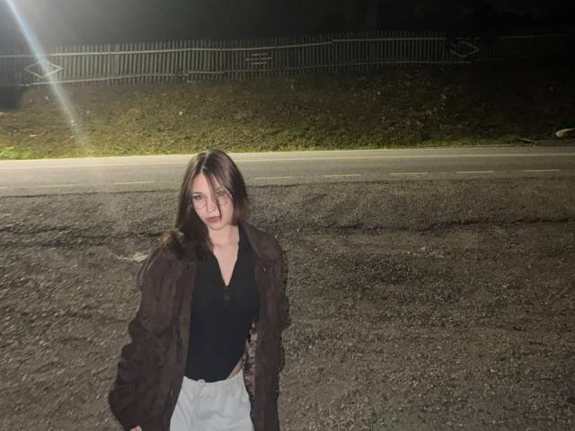 AletaBreslauer with long brown hair, black top, white pants, and brown jacket, standing on a gravel road at night, looking slightly annoyed.