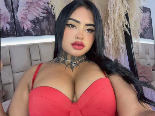 LairaHearvy with black hair, red bra, large boobs, tattoo on neck, choker, and red lipstick, looking at camera. Bedroom background with fluffy decor.