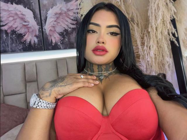 LairaHearvy with big tits in a red strapless top, black hair, tattooed neck, pink lips, and a silver watch, touching her chest. Bedroom with puffy paintings in background.