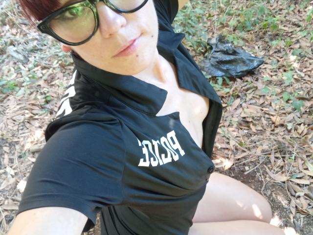 OrianaLaFrancaise with glasses, red hair, black shirt open to show tits, sitting on forest floor, legs apart, black leaf near her. She's looking at the camera.