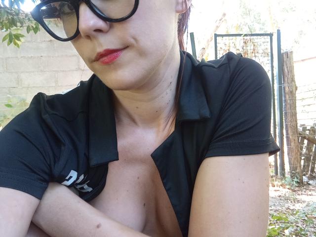 OrianaLaFrancaise with glasses, black shirt, and exposed boobs, arms crossed, red lipstick, light skin, and a slight smile, in a sunny outdoor setting.