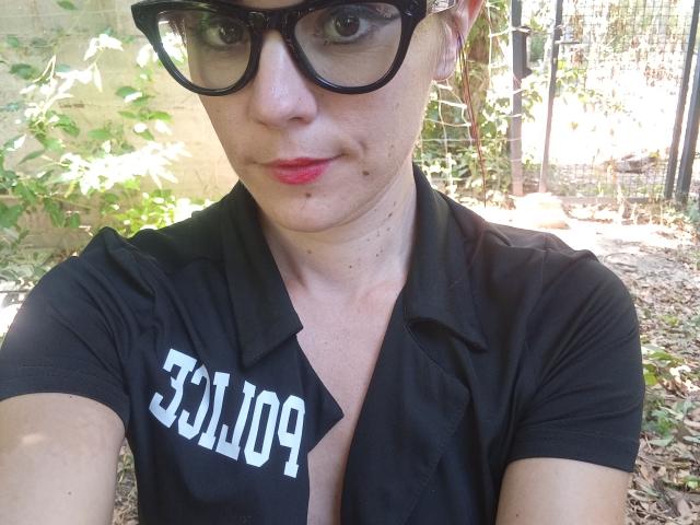 OrianaLaFrancaise with short brown hair, black glasses, red lipstick, black shirt, visible cleavage, black text, small zit on cheek, outdoors, daytime, green leaves in background.